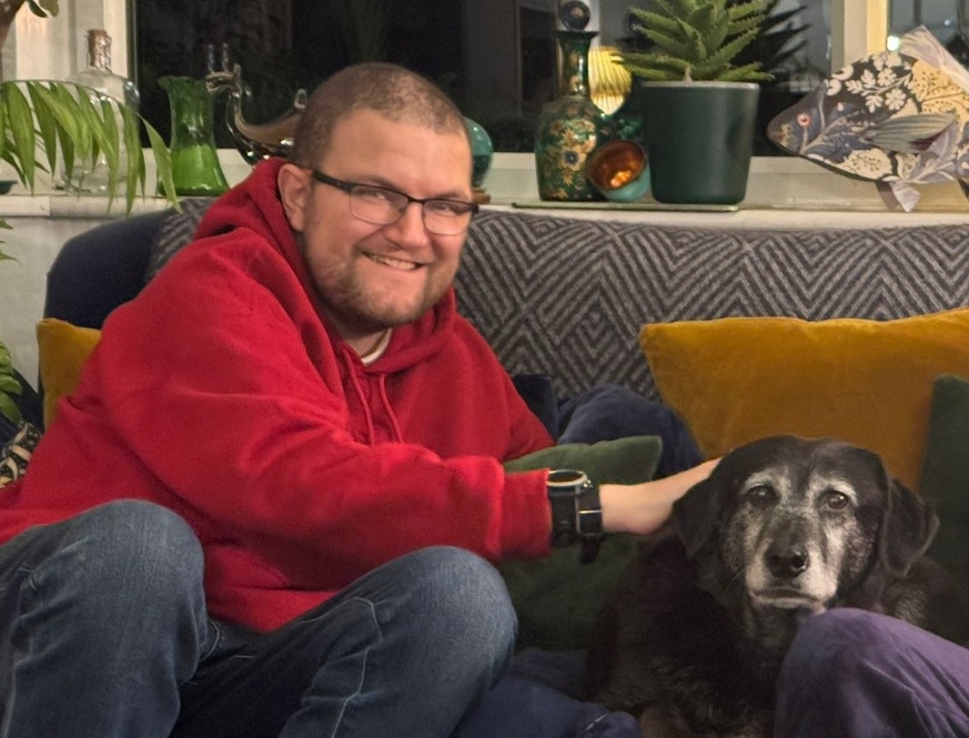 A picture of me wearing a red hoodie and glasses stroking my dog, Luka, large black dog with a white muzzle all around his nose. 