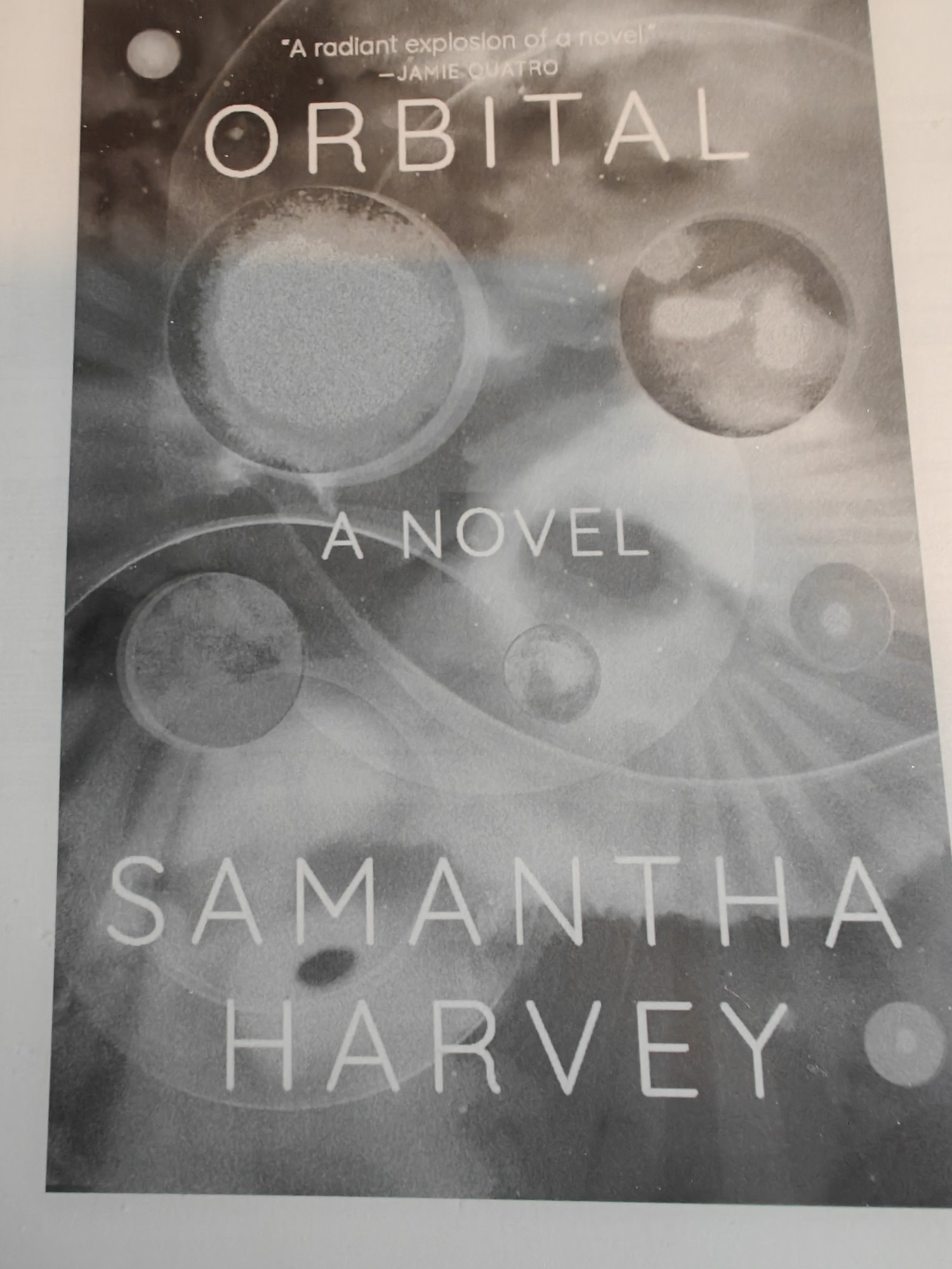 The front cover of Orbital by Samantha Harvey. It features some planets swirling about. My e-reader copy is black and white. 