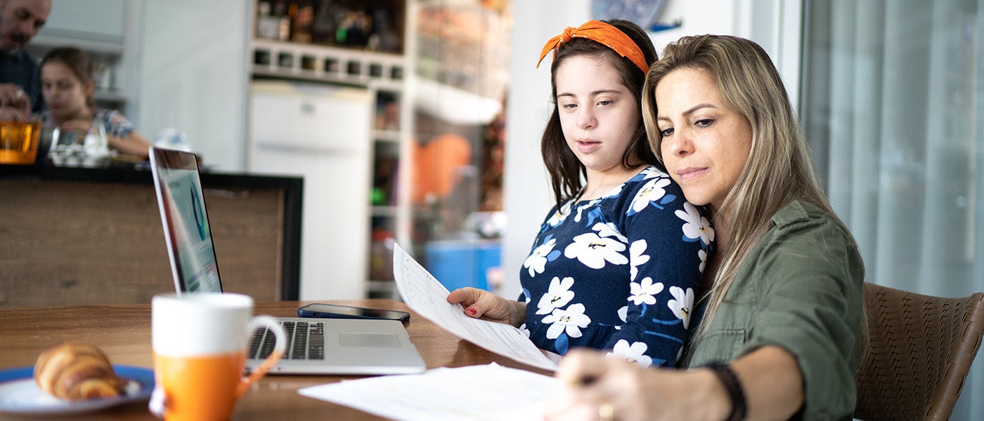 A picture of a girl with Down Syndrome with a woman, presumably her mother, sitting at a laptop, holding bits of paper