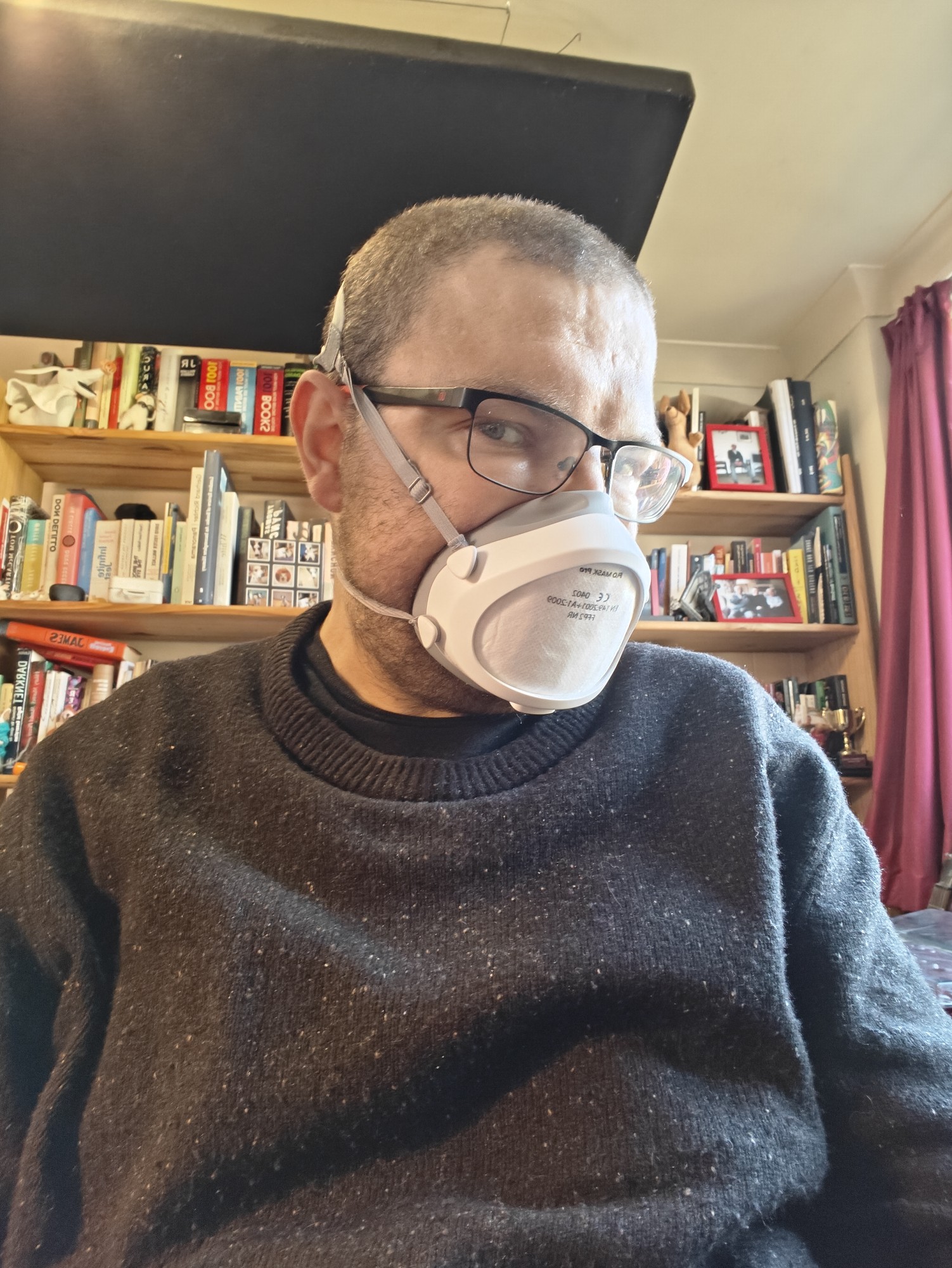 A picture of me, a bearded white man with very short hair and wearing a black speckled jumper, also wearing a Flo Mask respirator. 