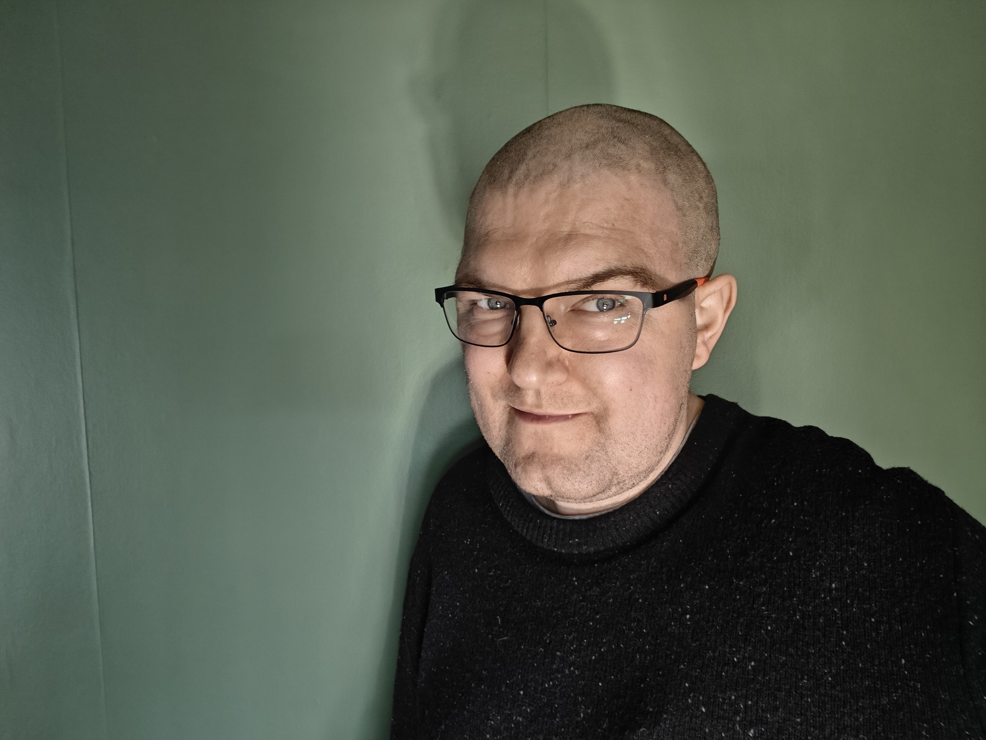 Me - a 40 year old man - wearing glasses with a black jumper against a green background. I now have no beard and only very very closely cropped hair. 