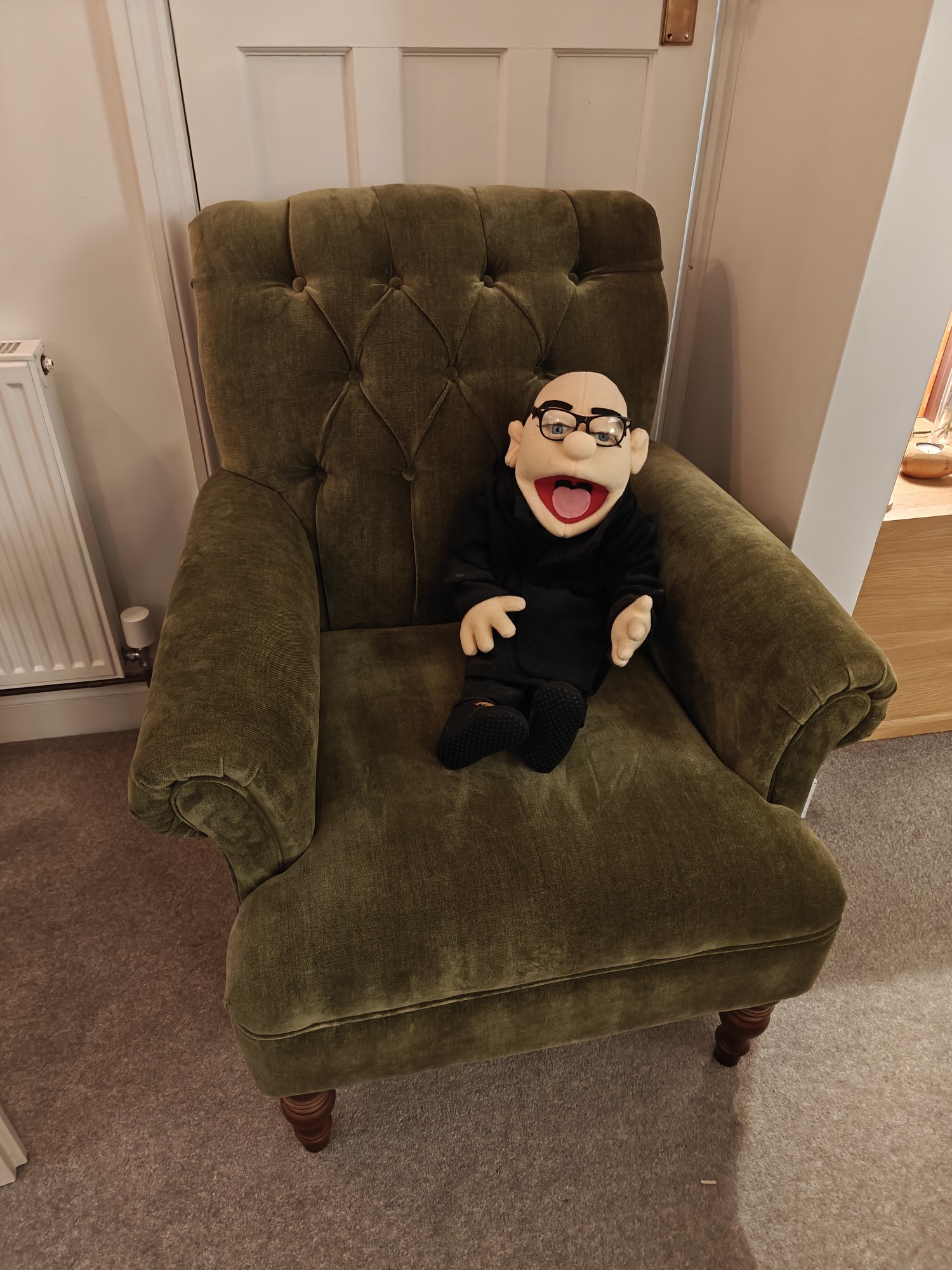 A green armchair with a puppet version of me in it. The puppet is bald/shaved head with spectacles, wearing black clothes. 