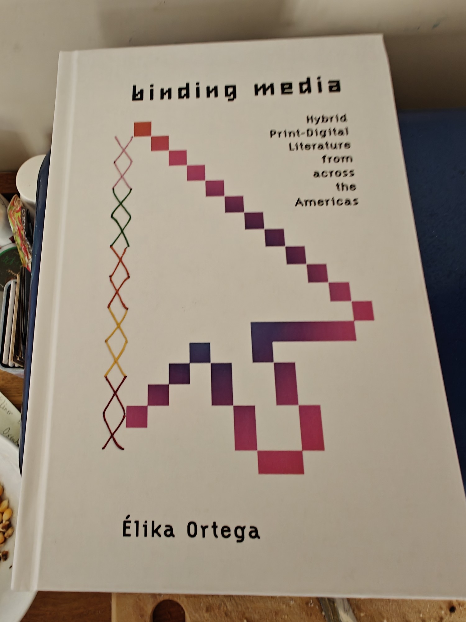 The cover of BINDING MEDIA: HYBRID PRINT-DIGITAL LITERATURE FROM ACROSS THE AMERICAS. It features a stylised mouse pointer. 