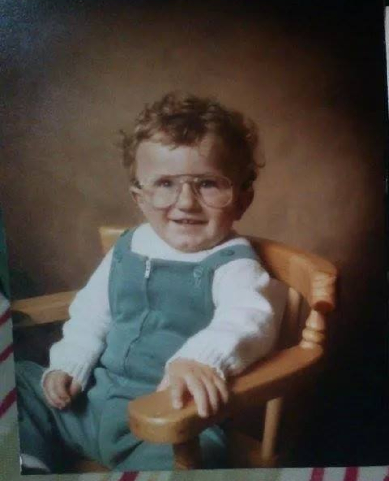 Me as a child with curly hair, sitting in a chair wearing dungarees
