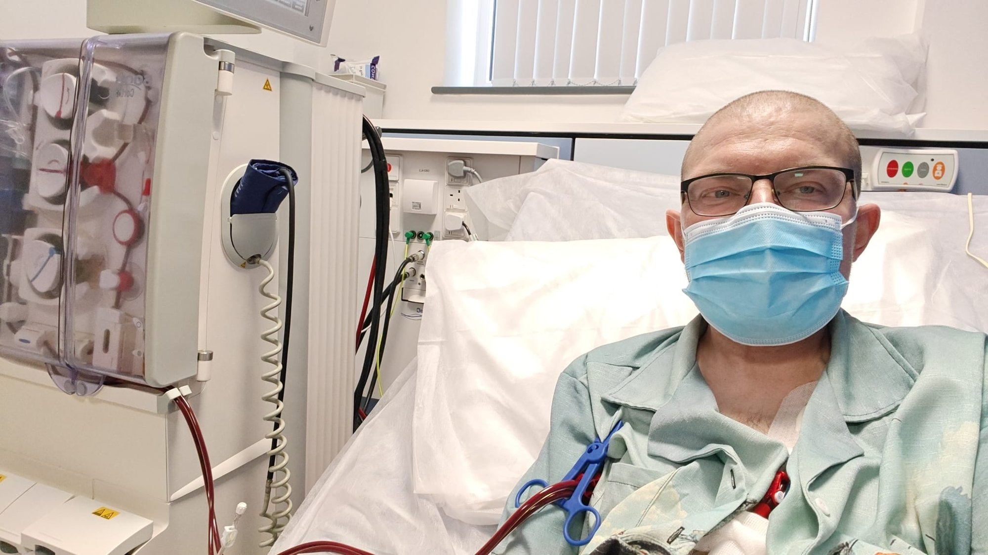 Me at the Bridgwater Dialysis Satellite Unit, wearing a surgical mask and a light bluey green shirt, with tubes coming out of my chest, attached to the dialysis machine to my left (photo is mirror image, so looks like it's on my right). 
