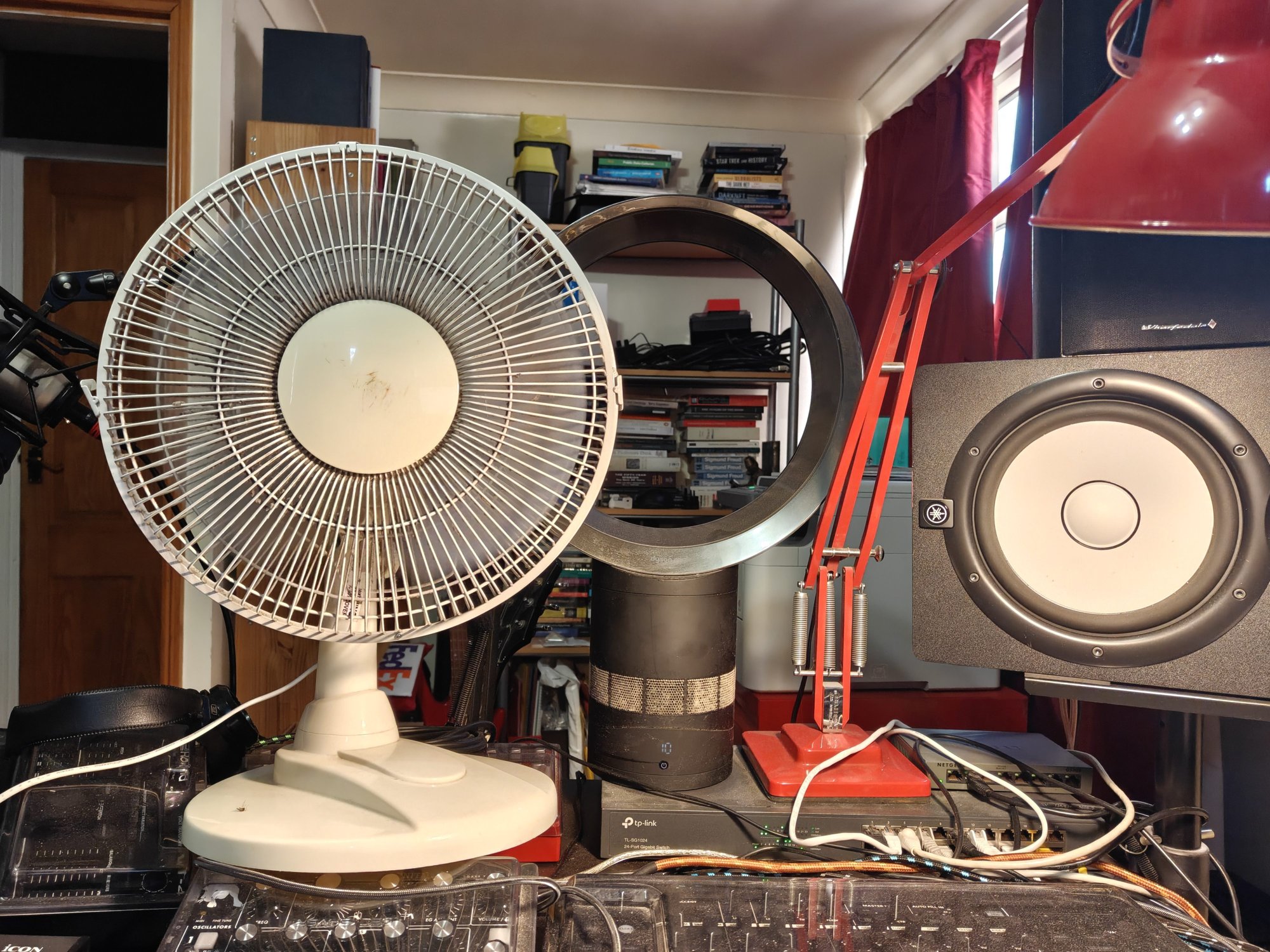 Two fans, one a Dyson bladeless, a red anglepoise lamp, and a large monitor speaker, all sitting atop some audio production equipment 