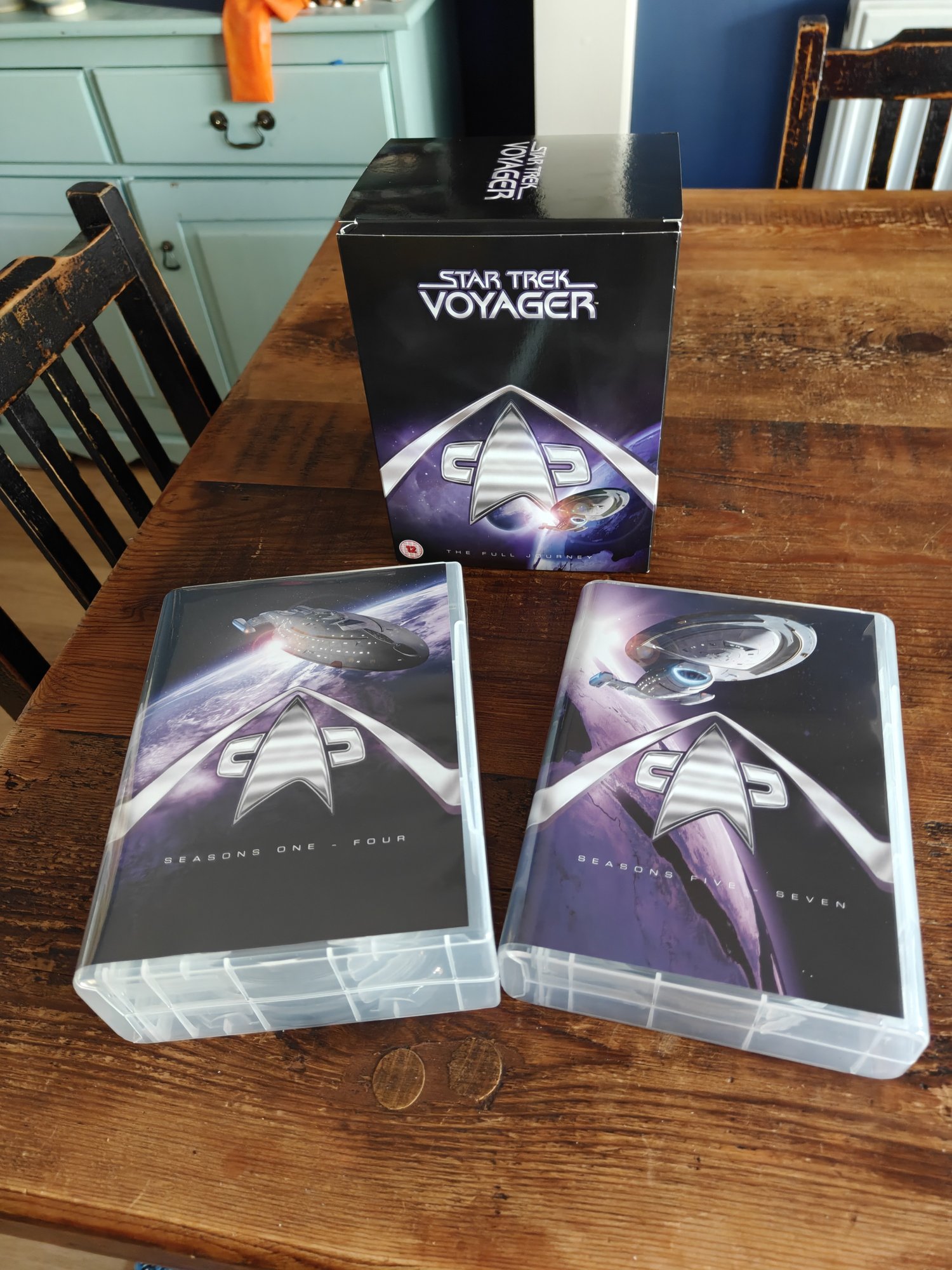 The DVD boxset of Star Trek: Voyager, with the box at the back and then the boxes for the discs laid out. They all feature Voyager, a starfleet insignia/badge, and the color scheme is black and purple.