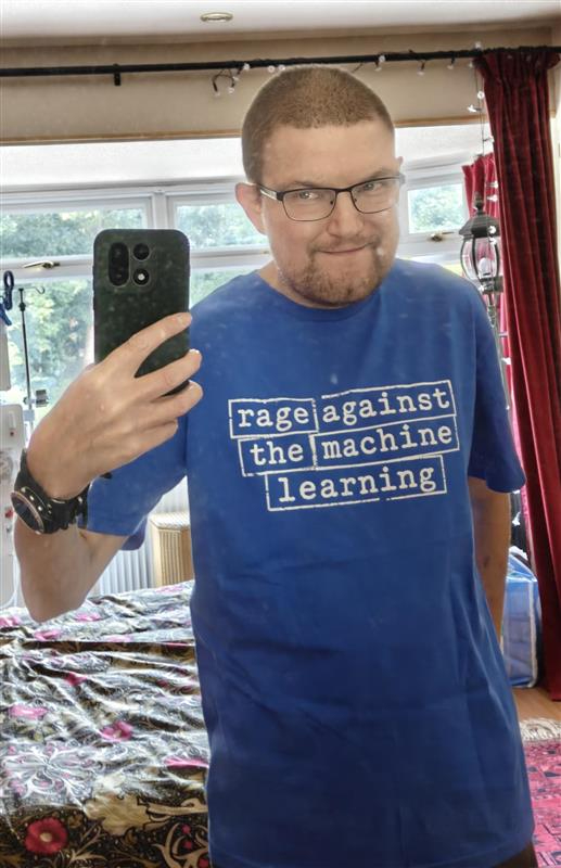 A photograph of me taking a selfie with the phone held up. I am a 6 foor 2 inch tall white male with short hair and glasses. I am wearing a blue t-shirt that says RAGE AGAINST THE MACHINE LEARNING in the logo style of the band Rage Against the Machine.