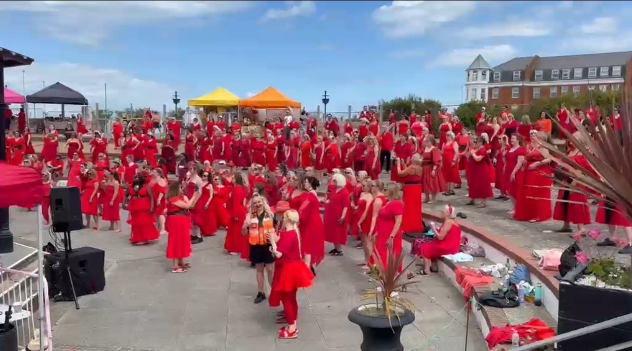 A large outdoor gathering of people dressed almost entirely in red fills a seafront plaza for a mass performance inspired by Kate Bush's Wuthering Heights. Hundreds of participants, many wearing flowing red dresses, skirts, or tops, are spread across stepped terraces and open paving, chatting and preparing to dance. In the foreground, a marshal in a high-visibility orange vest stands among the crowd, while speakers, barriers, and potted plants frame the performance area. Bright yellow and orange pop-up gazebos sit behind the dancers, with a large brick building and blue sky with scattered clouds in the background. The atmosphere is festive and communal, capturing the scale and anticipation of the famous Kate Bush The Most Wuthering Heights Day Ever dance gathering.