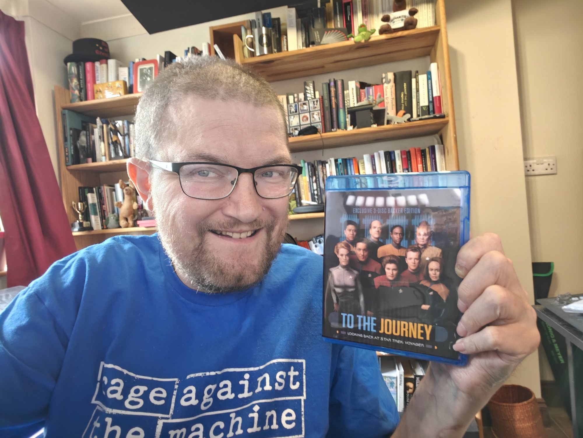 A smiling man - Martin Paul Eve, me! - with short hair, glasses, and a beard sits in front of floor-to-ceiling bookshelves filled with books and ornaments, holding up a Blu-ray case for the Star Trek: Voyager documentary To the Journey. They are wearing a bright blue Rage Against the Machine Learning T-shirt with the band's logo in white. The Blu-ray cover shows the main Voyager cast and reads "Exclusive 3-Disc Backer Edition" at the top and "To the Journey: Looking Back at Star Trek: Voyager" at the bottom. The room is warmly lit, with red curtains to one side, giving the image the feel of a personal library or study. 