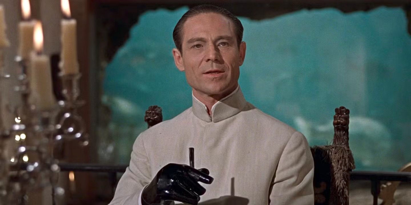 Dr. No from the 1962 James Bond film, 'Dr. No'. He is wearing a white up-collared shirt with black gloves on. There are large candles on the left of the image. The character is played by Joseph Wiseman, who, according to Wikipedia, is a "Canadian-born American actor of stage and screen". He is a white male, with a receding hairline but a strong black hair high on the forehead. He has no beard or mustache. He has a sinister air/look!